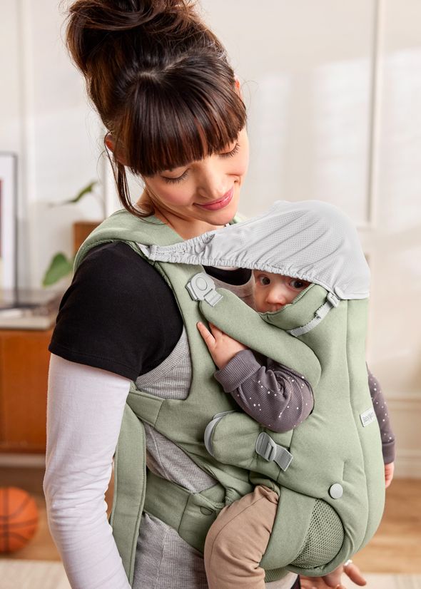 A woman smiles at a baby in a green carrier with a hood, as they stand in a cozy, sunlit room.
