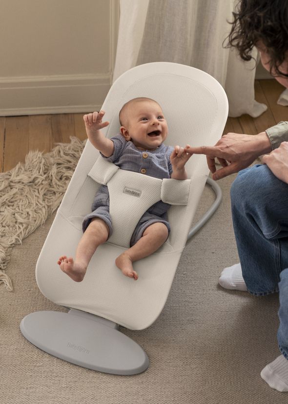 A baby in a gray outfit joyfully reaches for an adult's finger while sitting in a white BabyBjörn bouncer on a wooden floor.
