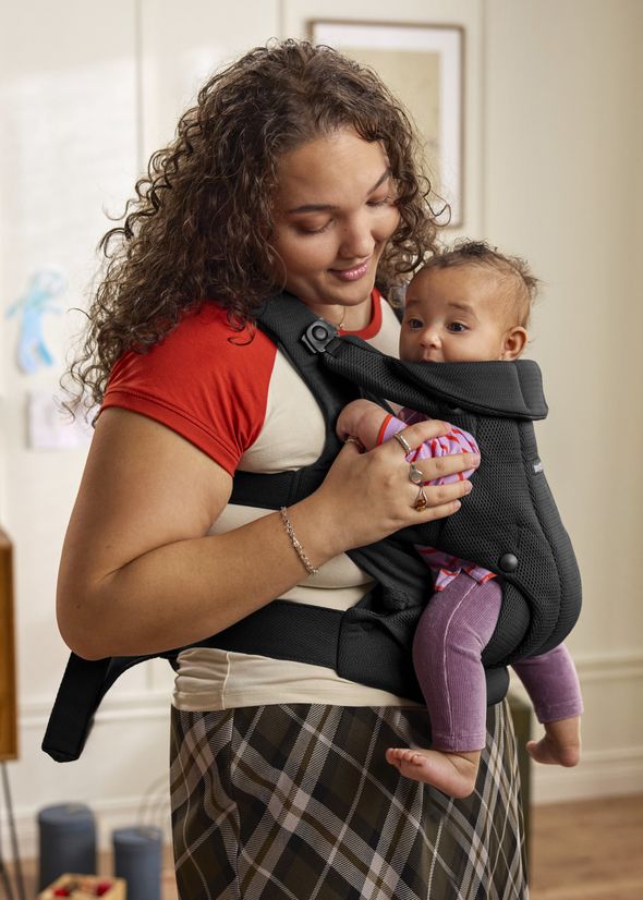 A woman in a red and white shirt holds a baby in a black carrier, smiling warmly. The baby wears pink leggings and looks content.