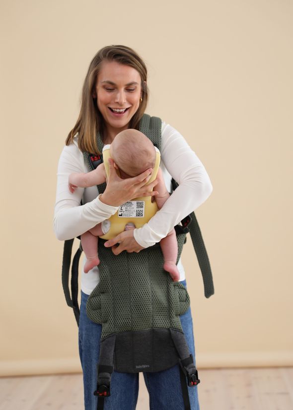 Woman smiling while adjusting a baby in a green carrier against a beige background
