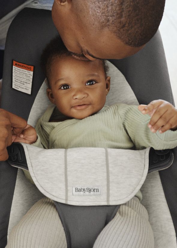 A baby sits in a BabyBjörn seat, smiling up at an adult who is gently kissing their head. The baby wears a light green outfit.