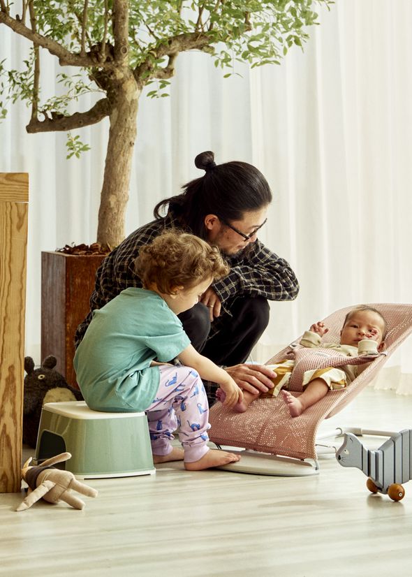 An adult and a child interact with a baby in a bouncer. The room has a potted tree, toys, and a wooden stool on a light-colored floor.