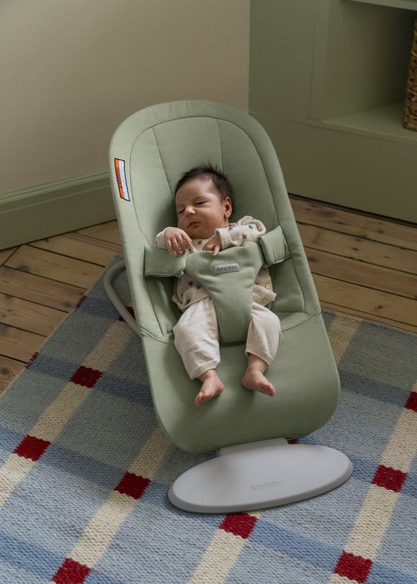 A baby sleeps in a green baby bouncer on a checkered rug, wearing a polka dot sweater and light pants.