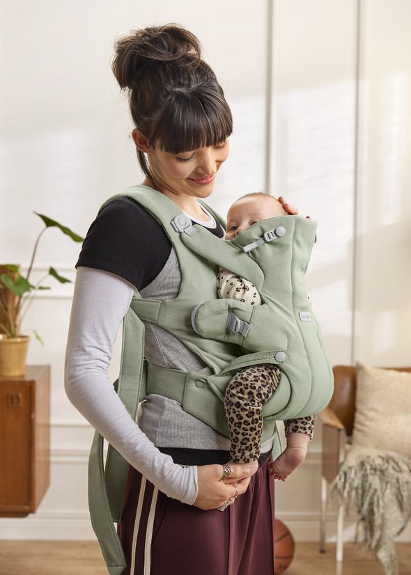 A smiling woman holds a baby in a green carrier, standing indoors. The baby wears leopard-patterned pants and a spotted shirt.
