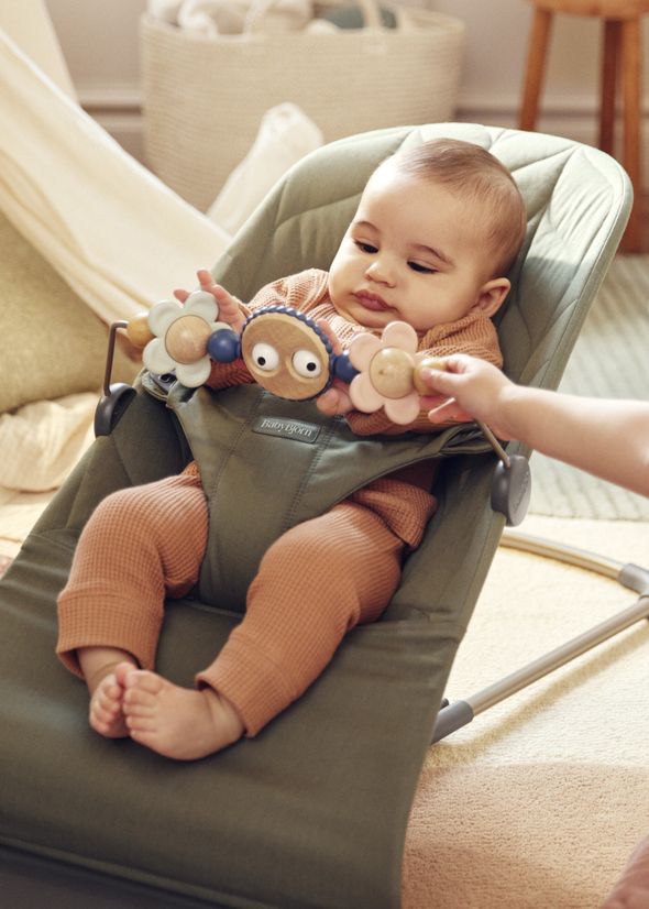 A baby in a green bouncer chair wearing a brown outfit, playing with a colorful toy bar with flowers and googly eyes.