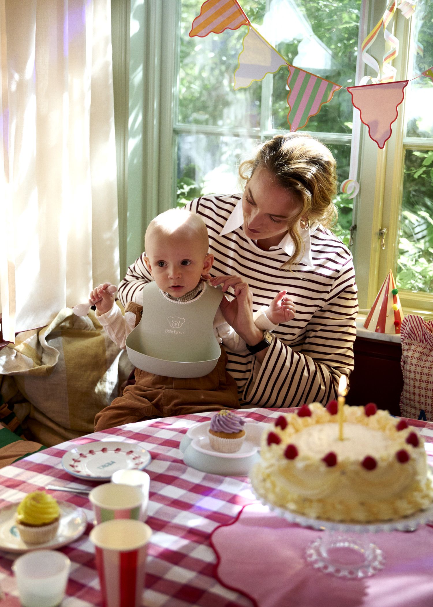 A woman holds a baby at a festive table with a cake and cupcakes. The room is decorated with colorful bunting and sunlight streams through the window.