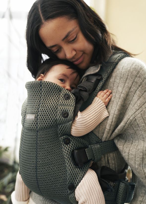 A woman smiles gently, cradling a baby in a green mesh baby carrier, both wearing cozy sweaters in a softly lit room.