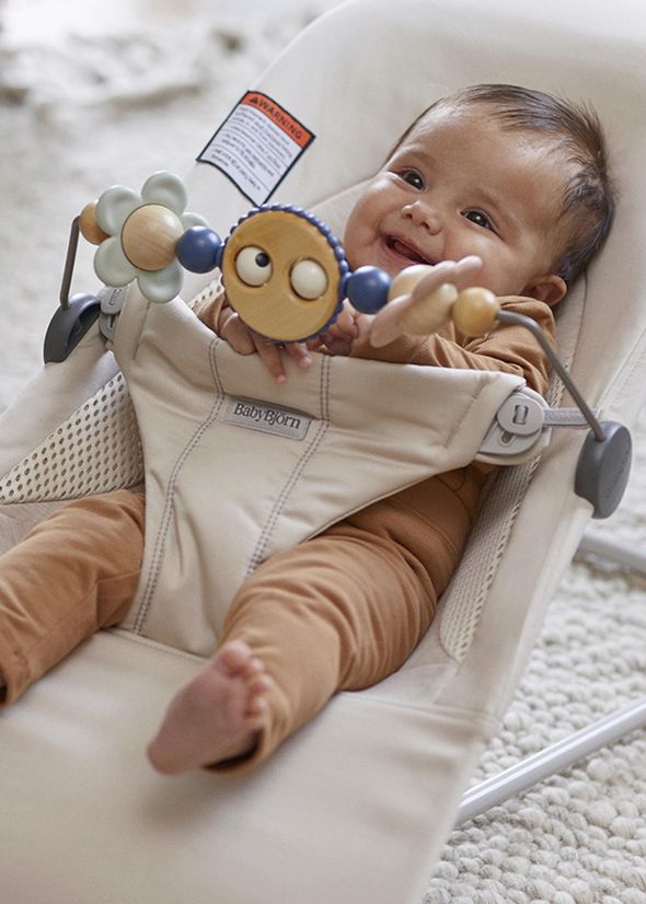 A baby in a beige onesie sits in a bouncer, smiling and holding a colorful toy with wooden rings and a flower-like design.
