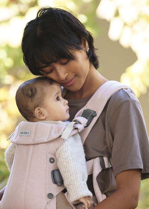A woman lovingly holds a baby in a pink carrier while standing outdoors, with soft focus trees in the background.