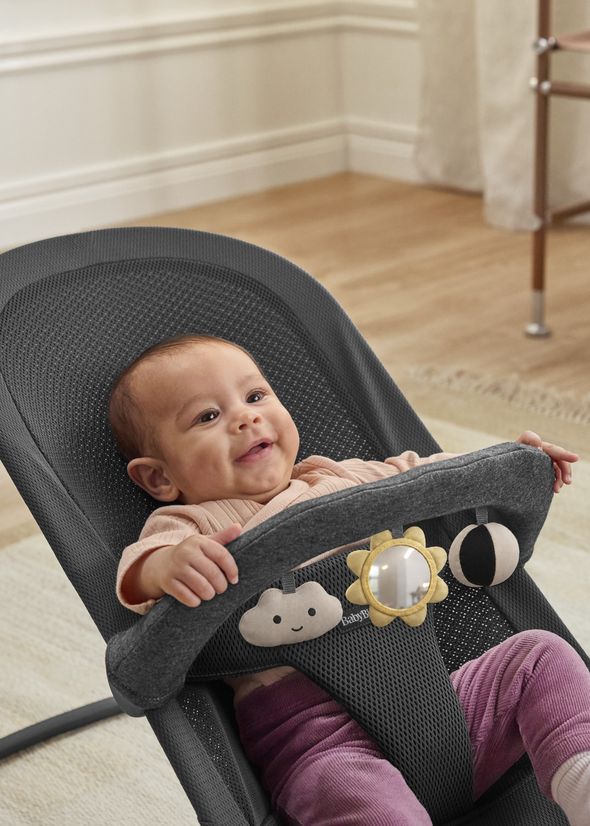 A smiling baby in a dark gray bouncer seat with toy attachments shaped like a cloud, sun, and ball. The baby wears a peach top and purple pants.