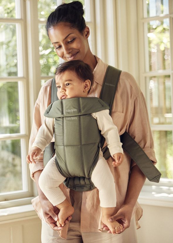 A woman smiles lovingly at a baby in a green baby carrier, standing by a window with natural light streaming in.