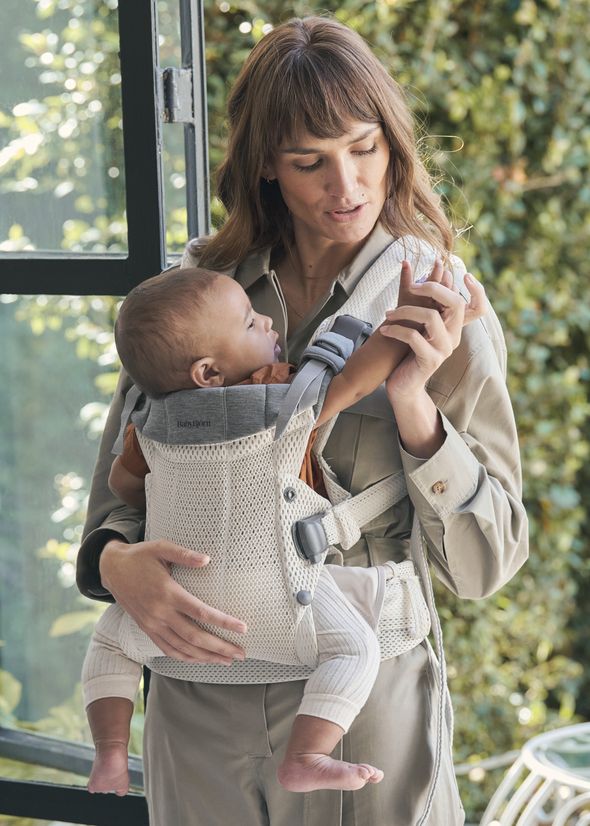 A woman holding a baby in a front-facing carrier, standing near a window with green foliage outside.