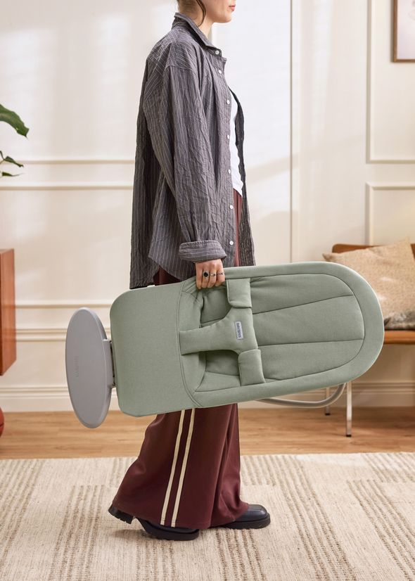 Person carrying a folded, light green baby bouncer indoors on a beige carpet, wearing a striped shirt and brown pants with stripes.
