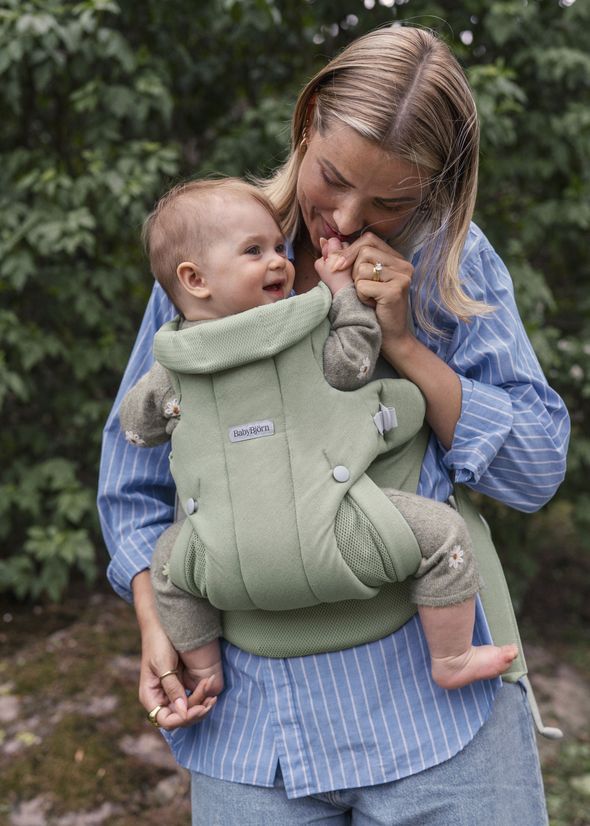 A smiling baby is carried in a green baby carrier by an adult outdoors. The baby is looking up, and the adult is wearing a blue striped shirt.