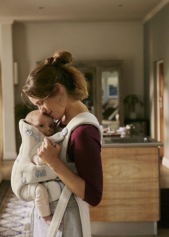 A woman in a maroon top gently holds and kisses a baby in a beige carrier inside a cozy, softly lit room.