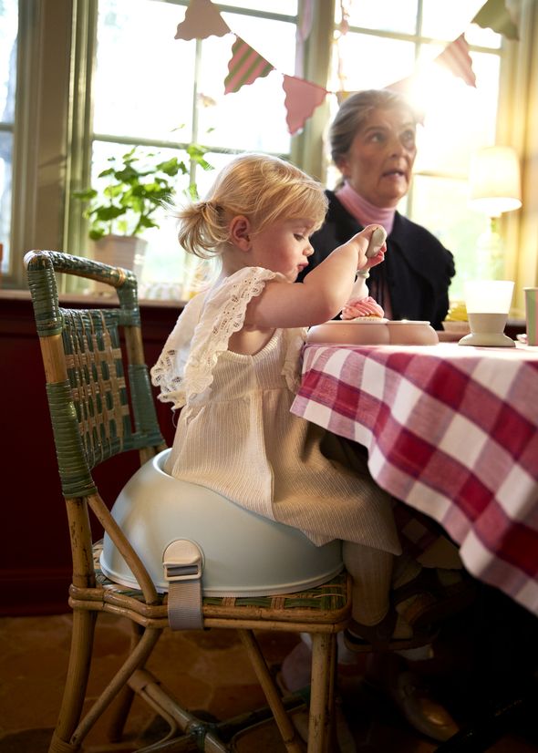 A toddler in a white dress sits on a chair eating at a table with a red checkered tablecloth, next to an older woman. Sunlight streams in.