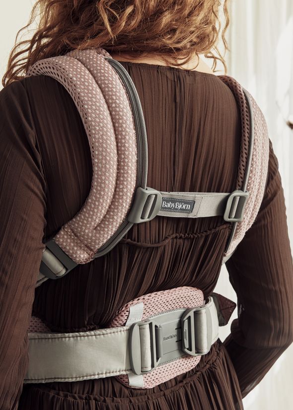 Person wearing a pink and gray BabyBjörn baby carrier over a brown dress, viewed from the back.