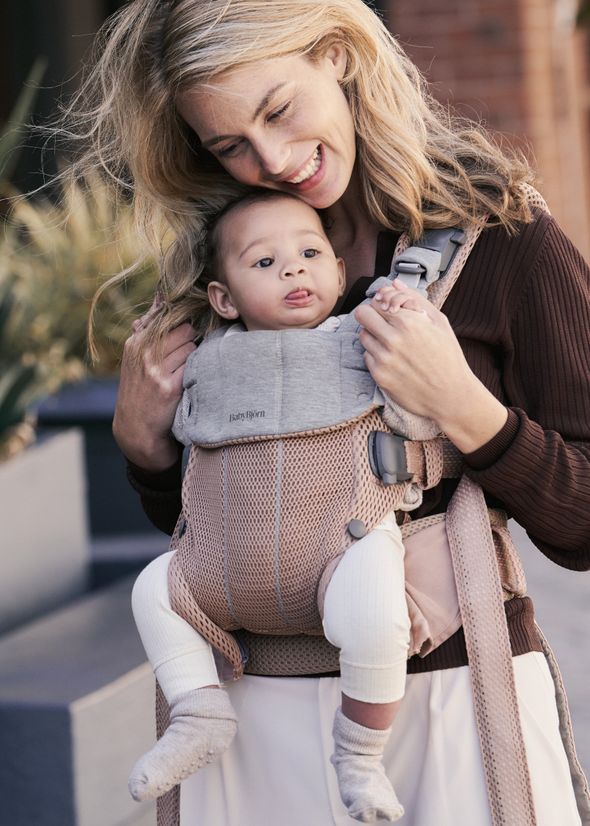 A woman smiles lovingly at a baby carried in a pink and gray front carrier, outdoors.