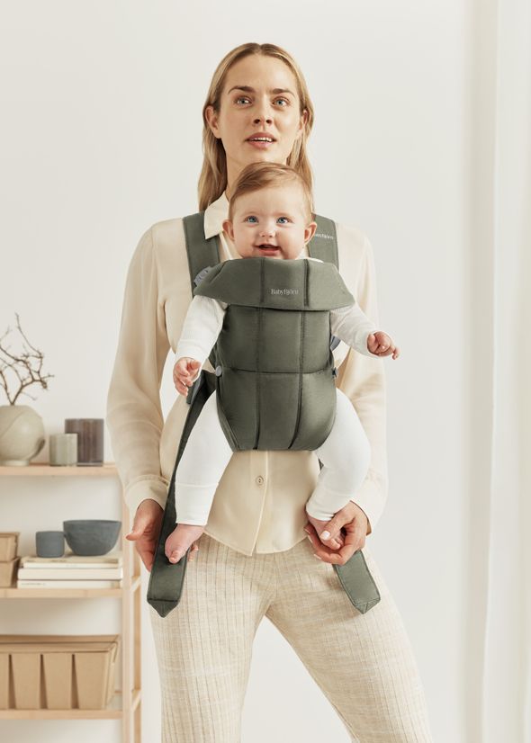 A woman wearing a light blouse and pants carrying a smiling baby in a green baby carrier. The background shows shelves with decor items.