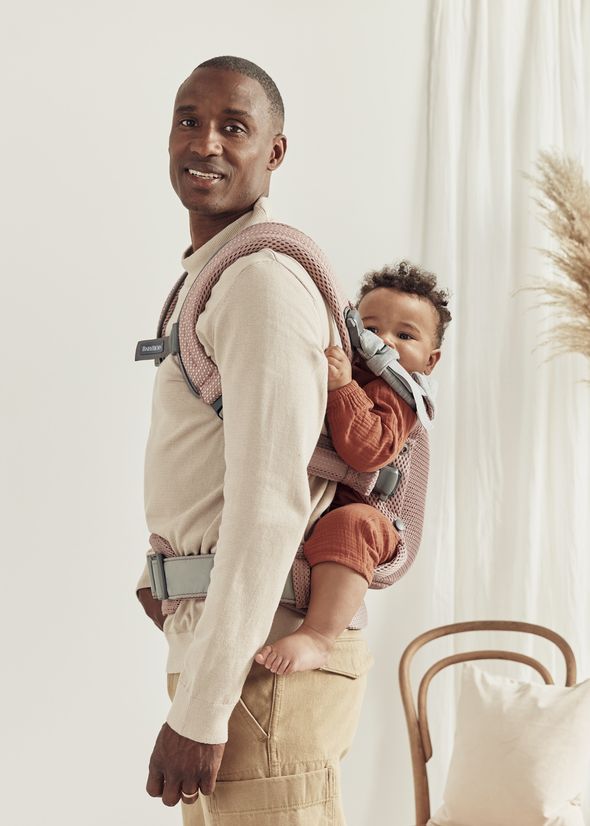 A man is smiling while carrying a baby in a pink baby carrier on his back. The baby is wearing a rust-colored outfit, and a chair is in the background.