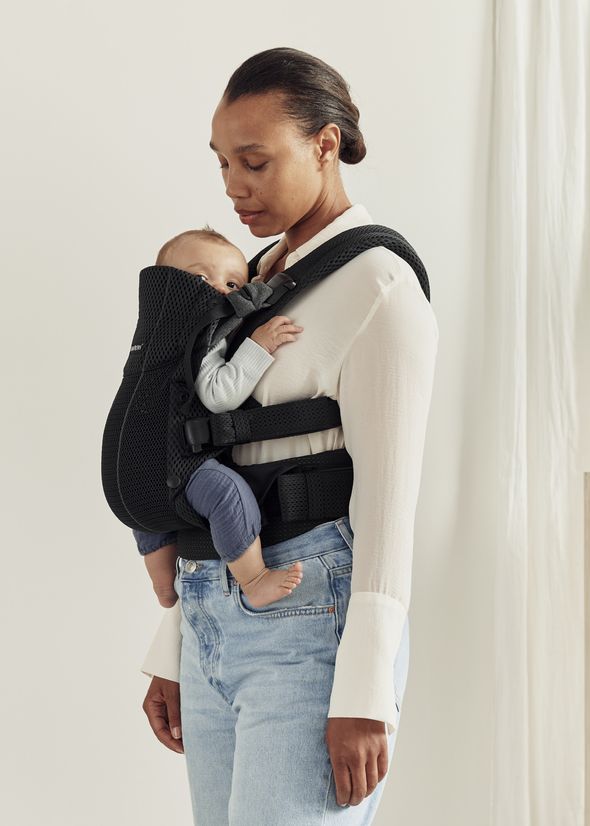 A woman stands indoors wearing a white blouse and jeans, holding a baby in a black front carrier. The baby is facing forward and wearing a blue outfit.