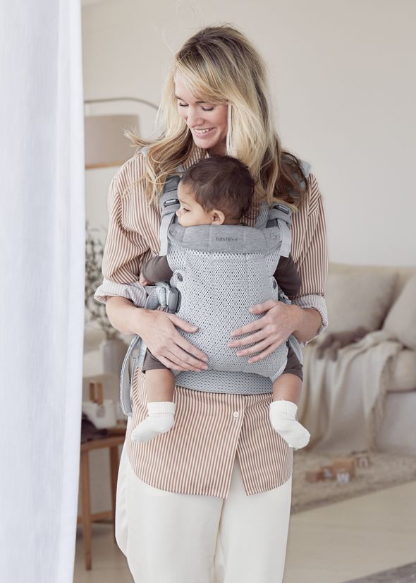 A woman smiles while holding a baby in a gray carrier in a cozy, softly-lit room with neutral colors.