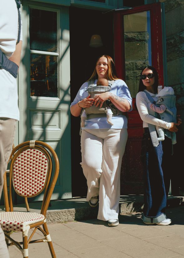 Two women with baby carriers stand outside a building; one is stepping out, and the other leans against the wall. A chair is in the foreground.