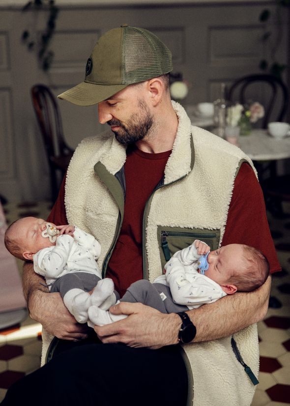 Man wearing a baseball cap holds two sleeping babies with pacifiers, dressed in matching outfits, in his arms indoors.