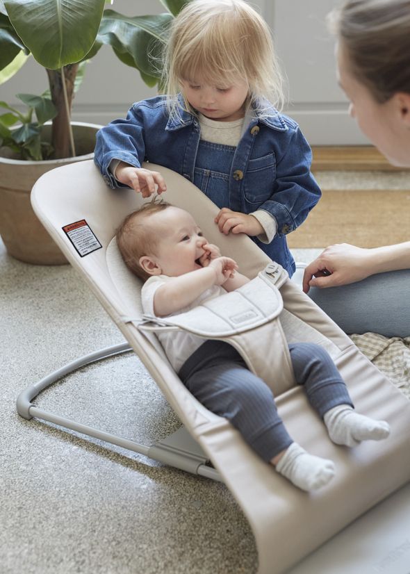 A toddler gently touches a baby lying in a beige baby bouncer, with an adult nearby. Indoor setting with plants in the background.