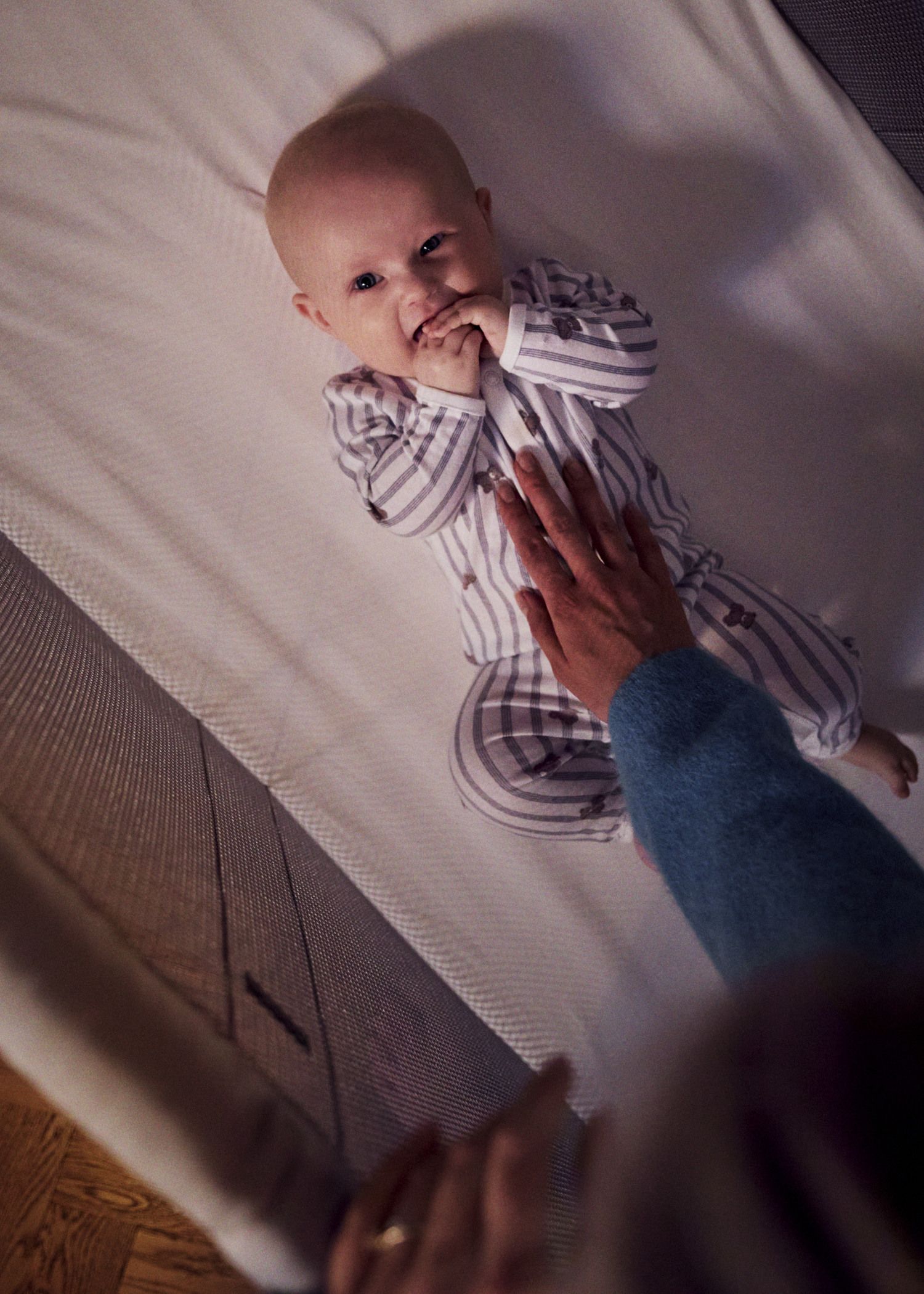 A baby in striped pajamas lies in a BabyBjörn Travel crib, smiling and touching its mouth, while a hand gently rests on its chest.