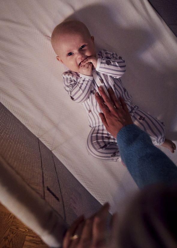 A baby in striped pajamas lies in a BabyBjörn Travel crib, smiling and touching its mouth, while a hand gently rests on its chest.