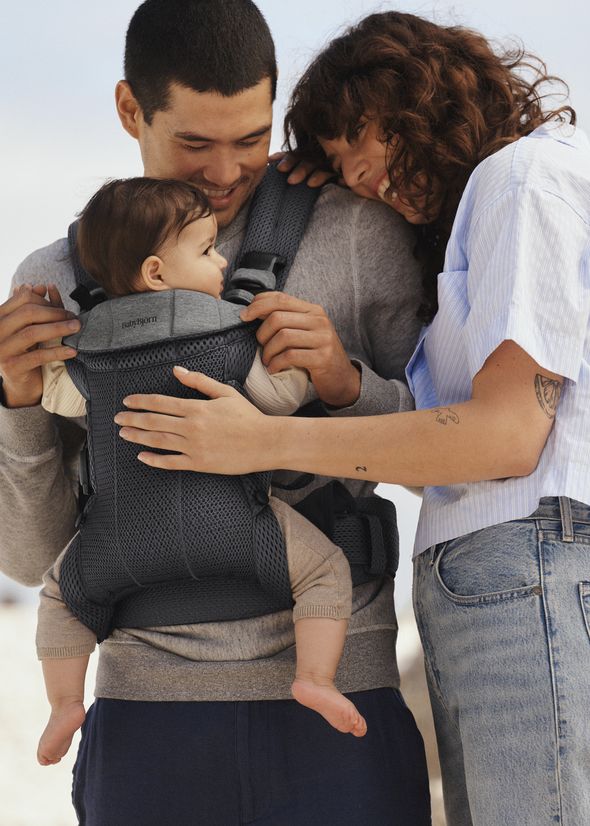 A smiling couple stands close, lovingly looking at their baby in a front carrier. The mother gently touches the baby's hand.