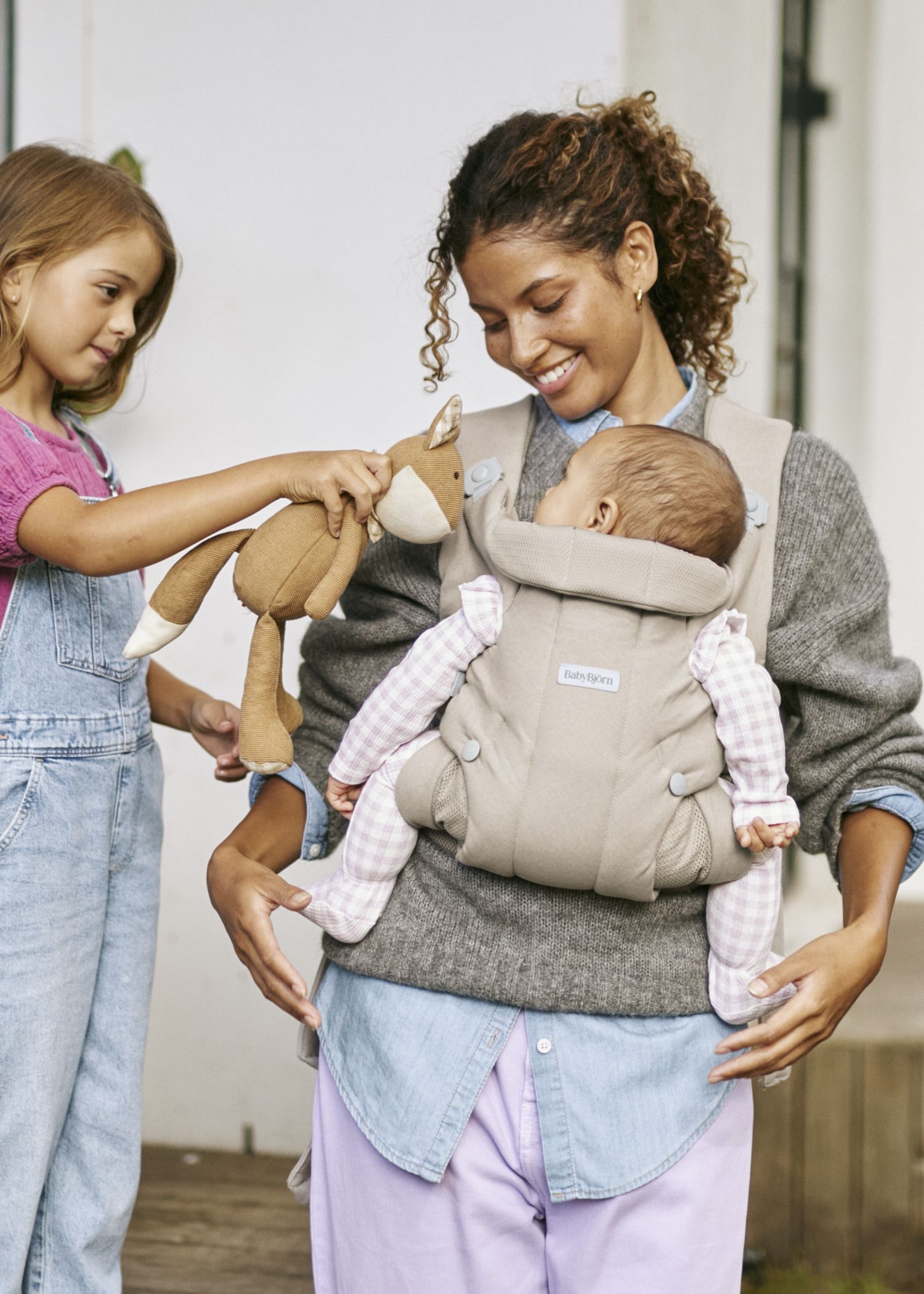 A woman smiles while holding a baby in a carrier. A young girl hands the baby a brown stuffed toy.
