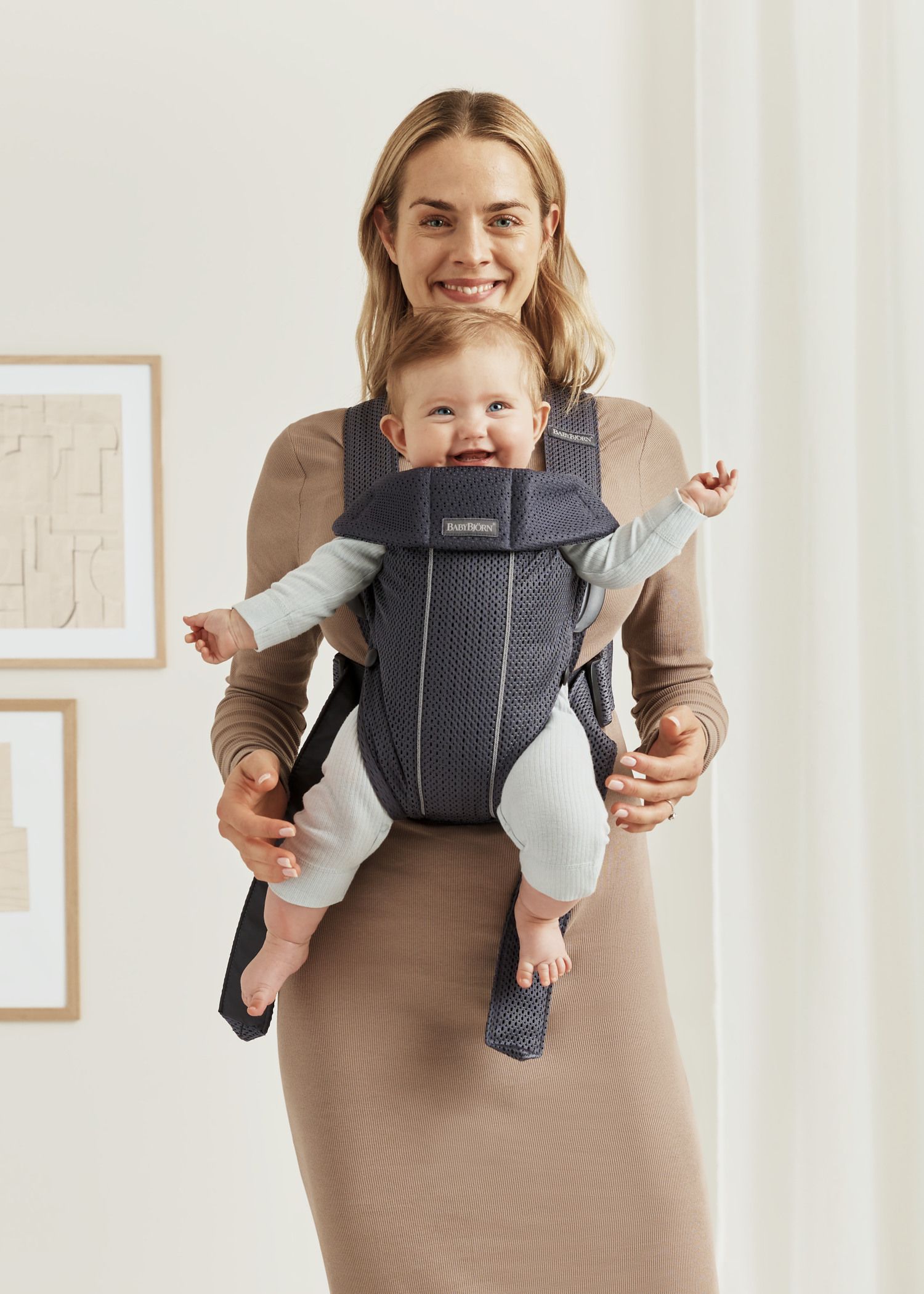 A smiling woman stands indoors holding a happy baby in a front-facing carrier. The room has framed art on the wall and soft lighting.