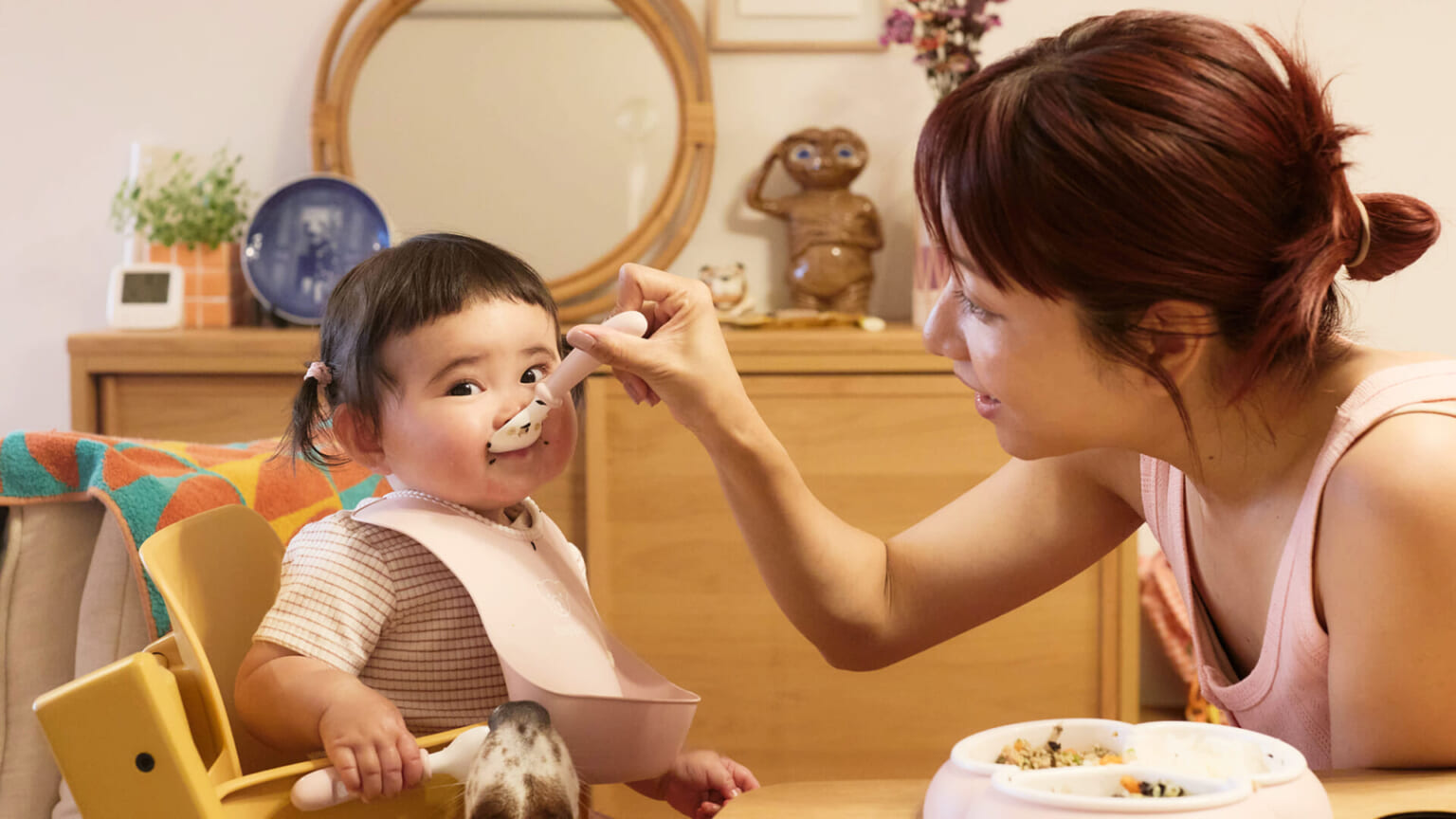 A woman feeds a smiling toddler in a high chair, while a dog looks up. A mirror and decorative items are in the background.
