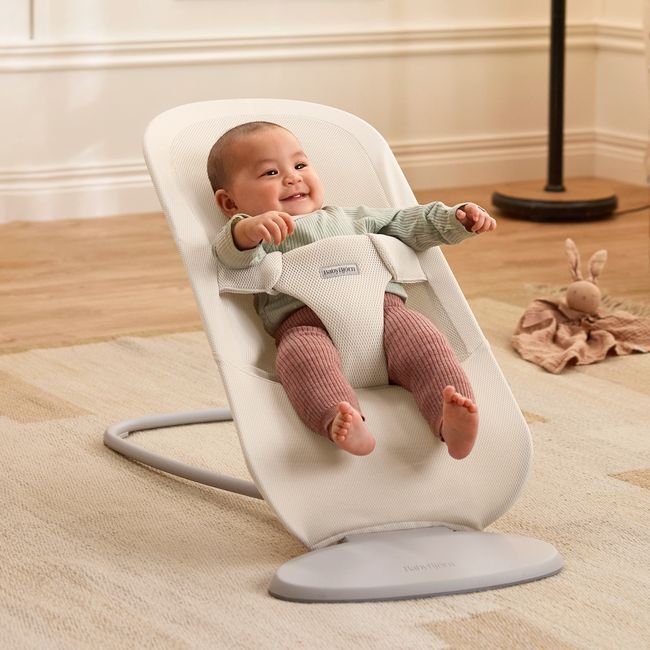 A smiling baby in a bouncer chair wearing a green shirt and pink pants, with a stuffed bunny toy on the floor nearby.