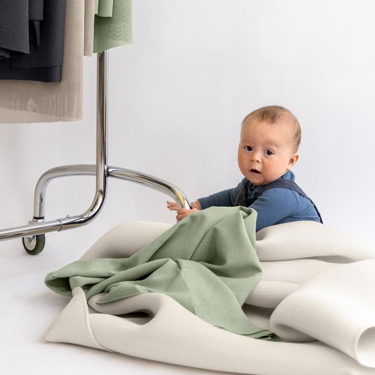 Baby in blue outfit sits on a floor surrounded by draped fabric, holding onto a garment rack in a well-lit area.