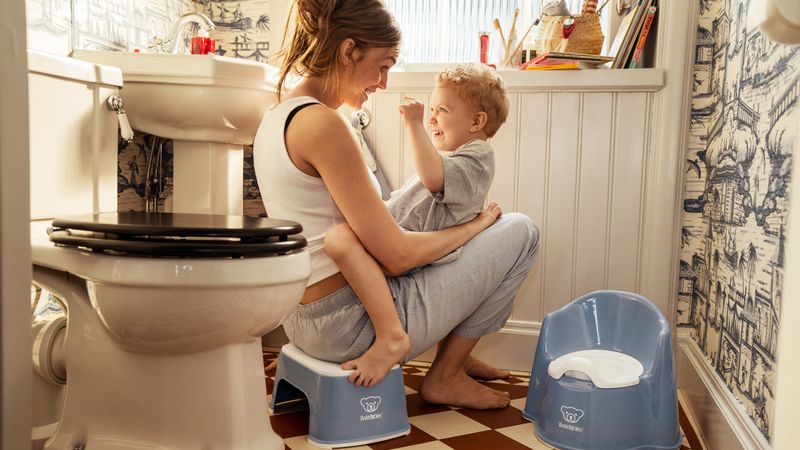 A woman and a child smiling at each other in a bathroom, with a potty chair and step stool nearby.