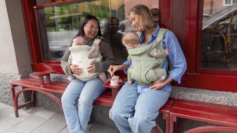 Two women seated on a red bench with babies in Baby Carrier Hold of Babybjorn , smiling and enjoying ice cream outside a storefront.