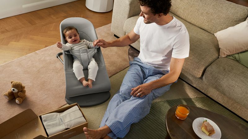A man in pajamas sits on a living room floor, holding hands with a smiling baby in a bouncer. A teddy bear and food are nearby.