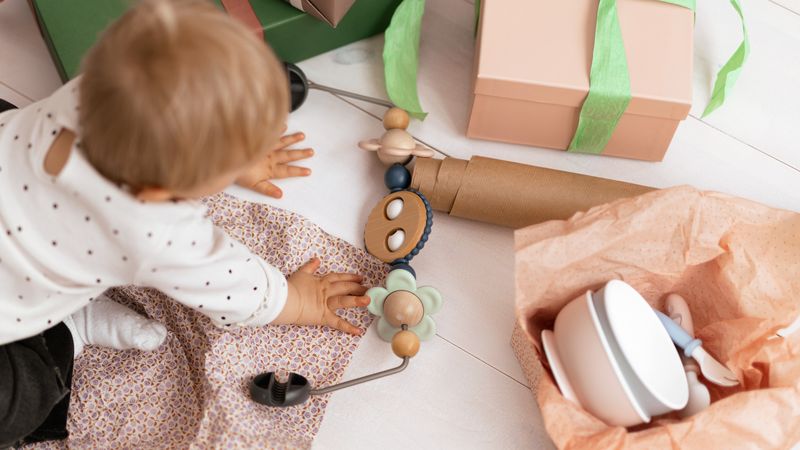 A baby in a polka-dot outfit reaches for wooden toys near gift boxes and a baby dish set wrapped in tissue paper on a light floor.