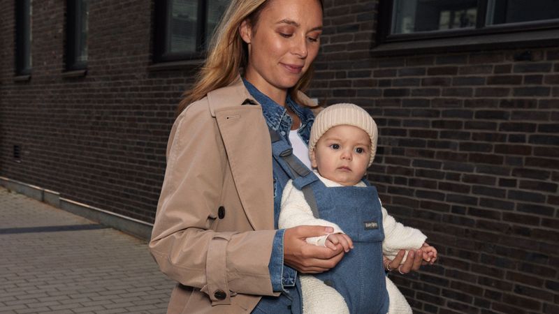 A woman walks outside holding a baby in a denim carrier. She wears a beige coat, and the baby is in a white outfit with a knit hat.