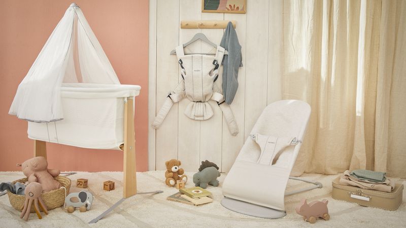 Cozy nursery with a white bassinet, baby carrier, bouncer, toys, and books. Neutral tones create a calm atmosphere with light curtains and soft carpeting.