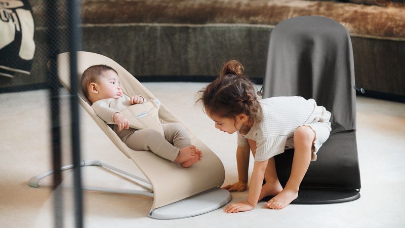 A toddler crouches beside a baby lying in a beige baby bouncer. Both are indoors on a light-colored floor.