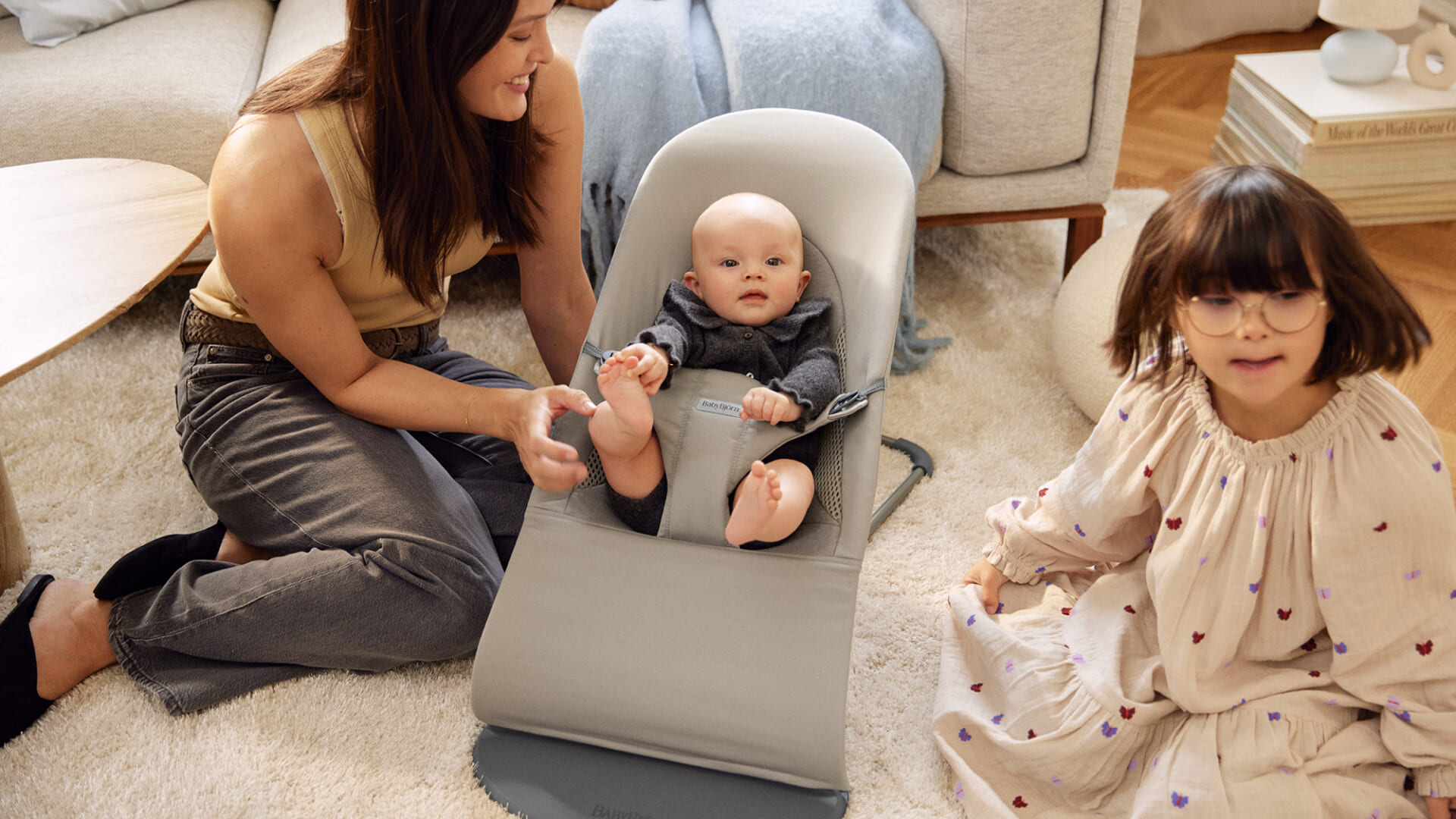 Bouncer Balance Soft in Light beige & Sand Grey Woven/Jersey Trifabric - BabyBjörn A woman sits on the floor with a baby in a bouncer and a young girl nearby, all in a cozy living room setting.
