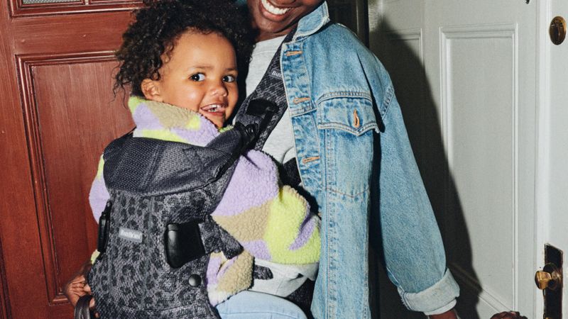 Smiling adult in denim jacket holding a cheerful child in a patterned carrier near a door.