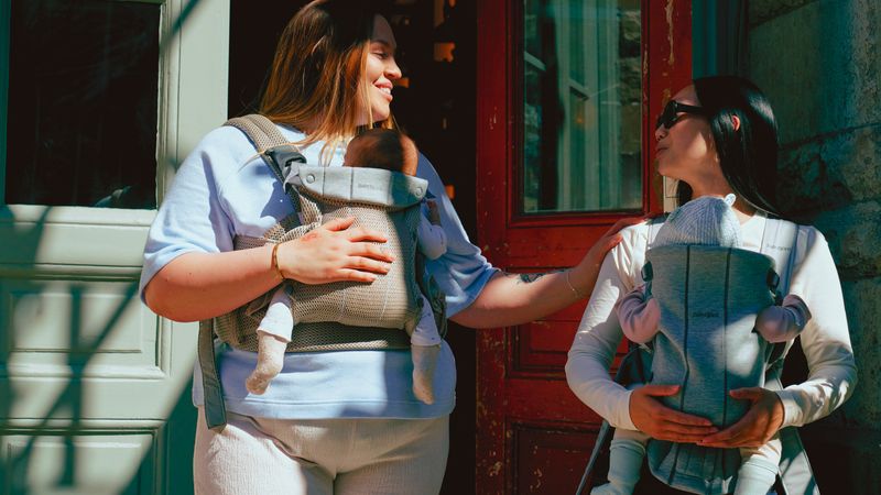 Two women smiling at each other, each carrying a baby in a front-facing carrier, standing outside a building with red and green doors.