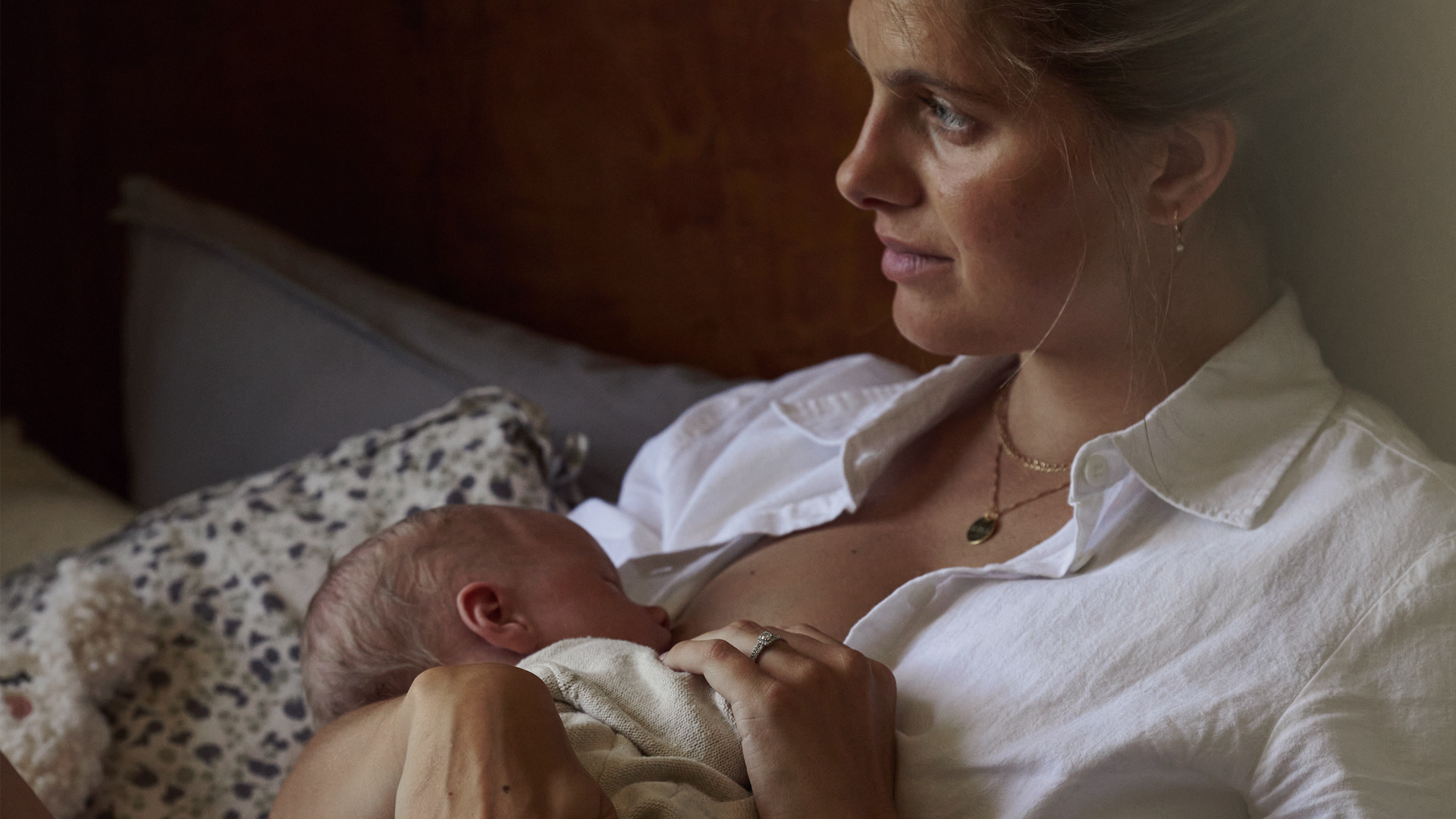A woman with a serene expression is breastfeeding her baby while sitting on a bed, wearing a white shirt and a gold necklace.