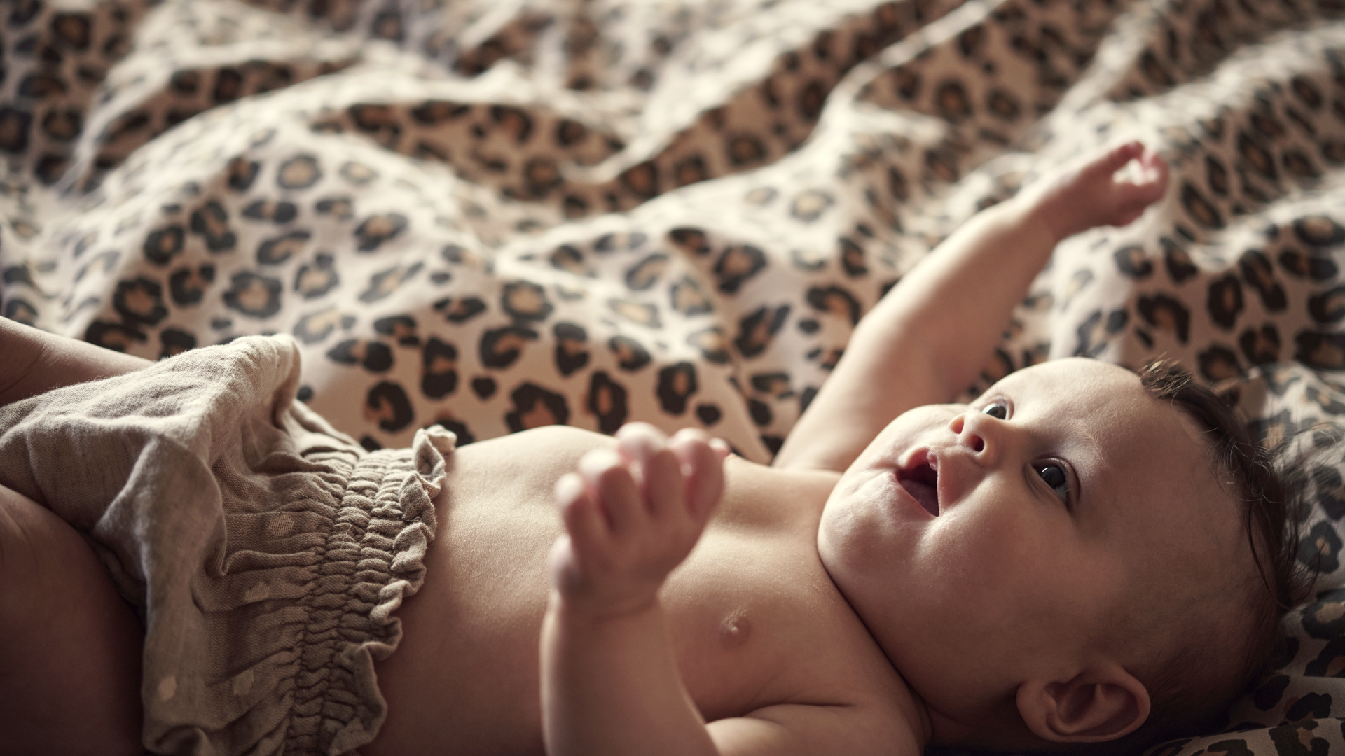 A smiling baby lying on a leopard-print blanket, wearing light-colored shorts, with arms raised and looking upward.