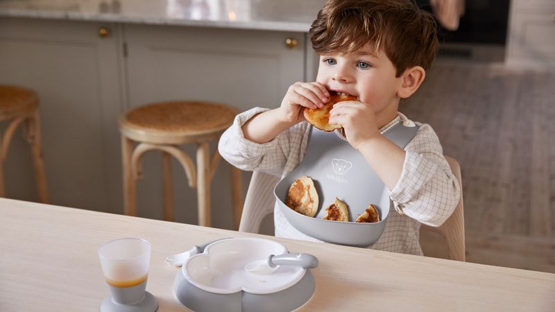 Young child eating a pastry at a table, wearing a bib. A plate and cup are in front of him on the wooden surface.